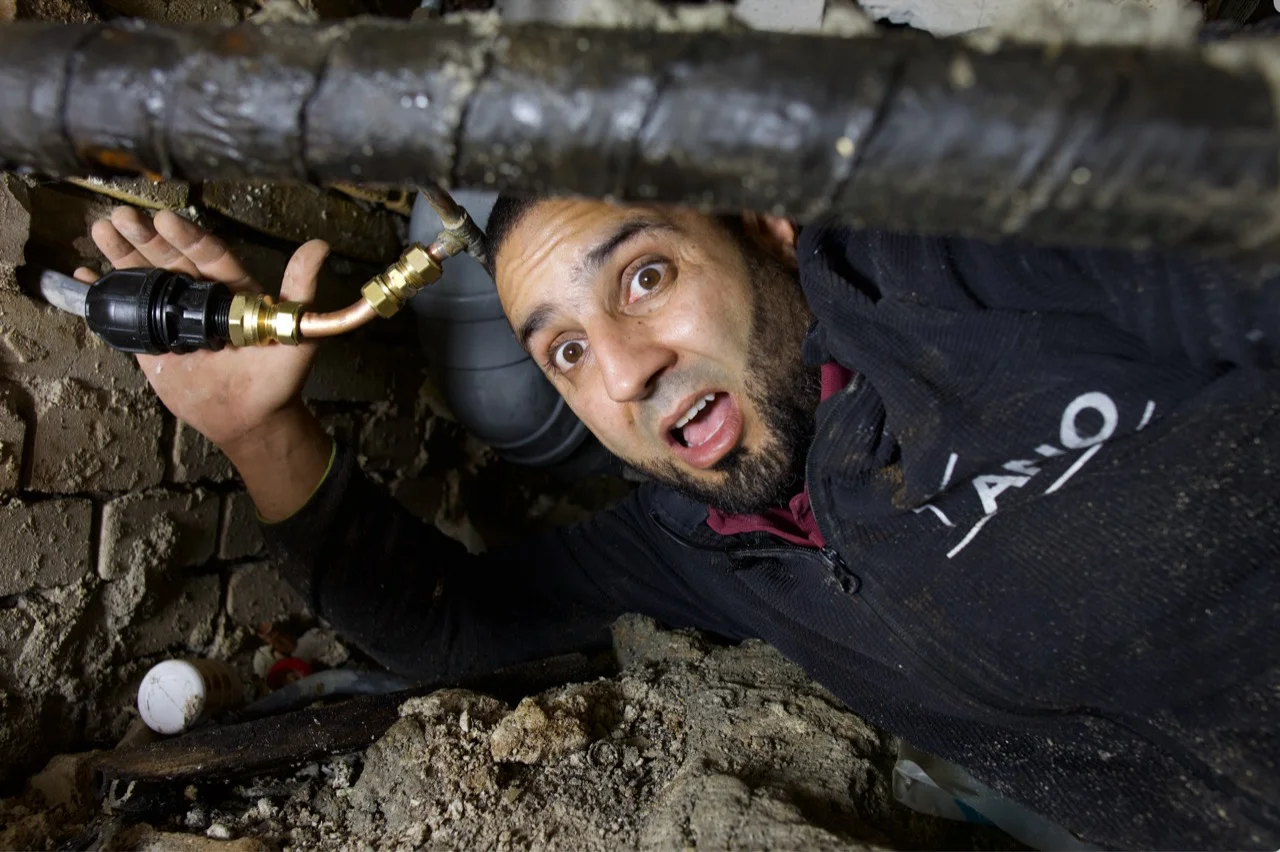 Loodgieter Anouar bij leidingwerk onder een vloer; gezicht, hand en aansluiting zichtbaar